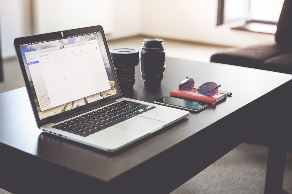 In a hotel room, a laptop on a table next to a phone, wallet, sunglasses and camera equipment.