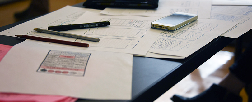 A desk covered in papers with drawing of smartphones, pens and an actual phone.