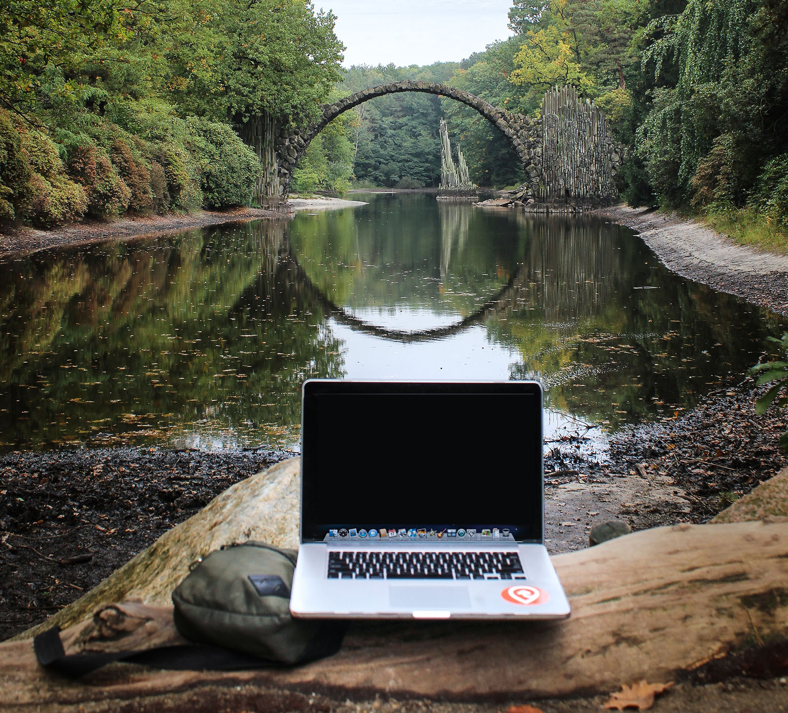 A laptop and backpack sitting on a log next to a river with a stone bridge in the distance.