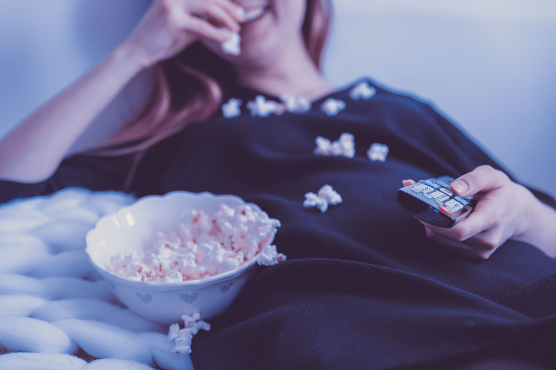 A woman laying down, eating popcorn with a remote in her hand.