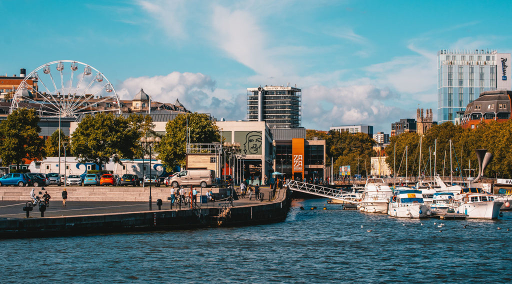 The city of Bristol, UK with people walking around a marina with boats and a Ferris wheel in the background.